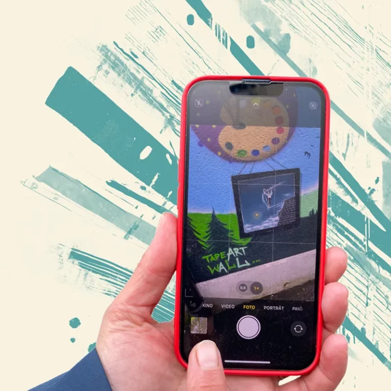 Smartphone in der Hand. Auf dem Display wird ein Fotomotiv einer bemalten Wand gezeigt, was gerade fotografiert werden soll.